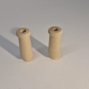 Puede incluir: Dos espigas de madera sin terminar. Cada espiga tiene un cuerpo cilíndrico y una parte superior ensanchada con un agujero. Las espigas son de color beige claro, lo que sugiere madera natural. Probablemente se utilizan en la fabricación de muebles o proyectos de artesanía.