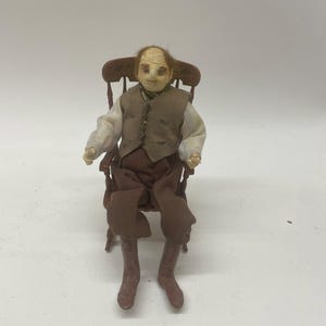Może przedstawiać: Miniaturowa lalka przedstawiająca starszego mężczyznę siedzącego na drewnianym krześle. Lalka ma na sobie białą koszulę, brązową kamizelkę i brązowe spodnie. Twarz lalki ma szczegółowe rysy i ma brązowe buty. Krzesło ma zakrzywione oparcie.