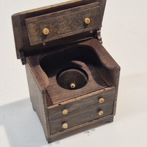 May include: A miniature wooden commode with a lift-up lid and two drawers. The commode is stained a dark brown color, with small, round knobs on the drawers and lid. The interior features a circular opening.