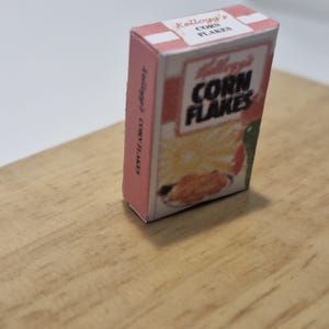 Op de afbeelding: Een miniatuur kartonnen doosje met Kellogg's Corn Flakes ontbijtgranen. De doos is roze en wit met een afbeelding van de ontbijtgranen op de voorkant. De tekst "Kellogg's Corn Flakes" staat op de doos gedrukt.