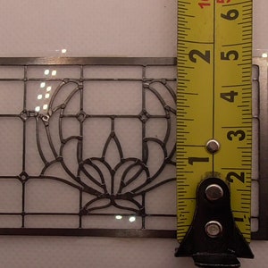 Op de afbeelding: Een transparante kunststof sjabloon met een zwart metalen frame, 7,6 cm hoog, met een bloemenmotief. De sjabloon wordt gemeten met een gele meetlint.