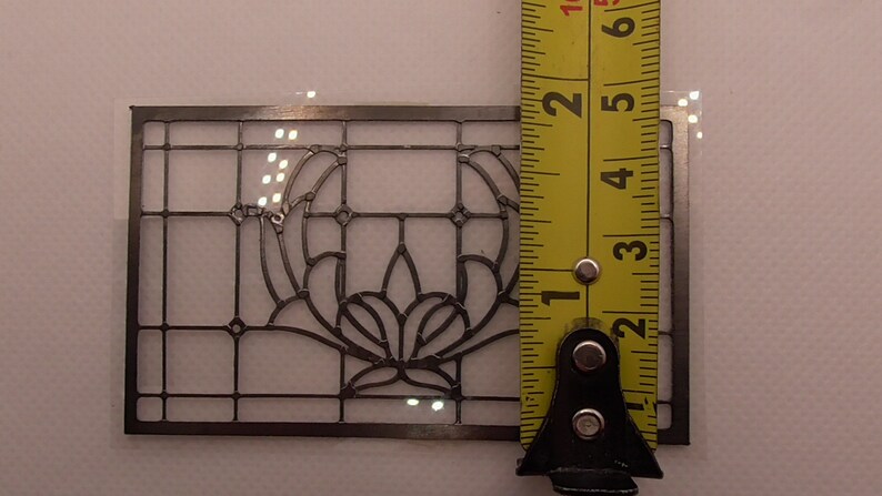Op de afbeelding: Een transparante kunststof sjabloon met een zwart metalen frame, 7,6 cm hoog, met een bloemenmotief. De sjabloon wordt gemeten met een gele meetlint.