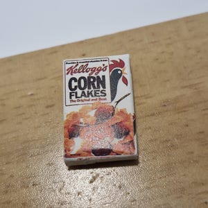Op de afbeelding: Een miniatuurdoos Kellogg's Corn Flakes ontbijtgranen. De doos is wit met rode en zwarte letters en een rode haanillustratie. De tekst op de doos luidt "Kellogg's Corn Flakes The Original and Best".