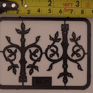 Könnte beinhalten: Ein schwarzer Kunststoff-Anguss mit zwei detaillierten Metallscharnier-Designs. Der Anguss ist mit "BCM" beschriftet.