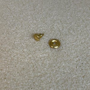 May include: A small, gold-colored earring stud and backing. The stud has a textured design, and the backing is a flat disc with a circular pattern. The items are displayed on a textured, off-white surface.