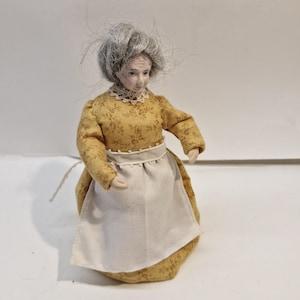 Peut inclure: Une poupée miniature représentant une personne âgée aux cheveux gris, vêtue d'une robe jaune moutarde à manches longues avec un petit motif floral marron, un col en dentelle blanche et un tablier blanc uni noué à la taille avec des boutons.