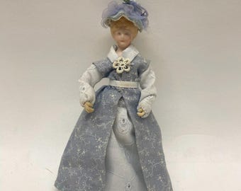May include: A porcelain doll dressed in a light blue and white patterned dress and matching hat. The doll has a white collar and a white belt. The doll's dress has a floral pattern. The doll is a miniature collectible.