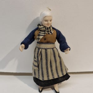 May include: A porcelain doll with white hair wearing a blue long-sleeved top, a brown bodice, and a beige apron featuring vertical black stripes. The doll is dressed in a black skirt and black shoes, with a matching black and white striped neck scarf.