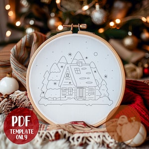 May include: A white embroidery hoop with a black line drawing of a winter cabin with snow-covered mountains and trees. The design is perfect for a cross-stitch or embroidery project.