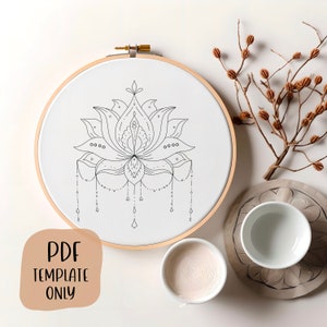 May include: A black and white line drawing of a lotus flower with a decorative border and hanging teardrop shapes. The drawing is on a white canvas in a wooden embroidery hoop.
