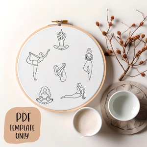May include: A white embroidery hoop with a wooden frame, featuring six black line drawings of women in various yoga poses. The hoop is on a white surface with a small white bowl and a branch with brown dried berries.