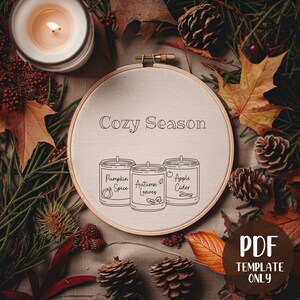 Peut inclure: Un cerceau de broderie beige avec le texte "Cozy Season" et trois bougies aux senteurs "Pumpkin Spice", "Autumn Leaves" et "Apple Cider".