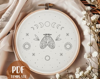 Può includere: Un cerchio da ricamo bianco con un disegno a linea nera di una falena, lune e soli. Il cerchio è su uno sfondo di tessuto bianco.