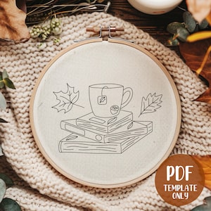 May include: A black and white line drawing of a cup of tea with a tea bag, a stack of books, and two leaves on a white background. The image is in a round embroidery hoop.