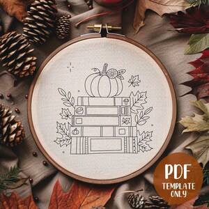 May include: A black and white line drawing of a stack of books with a pumpkin on top. The books are decorated with fall leaves and a mushroom. The image is on a beige fabric background.