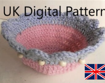 May include: A crocheted bowl with a pink and gray color scheme. The bowl has a scalloped edge and white beads around the rim.