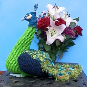 May include: A green and blue crocheted peacock figurine with a long neck and a tail made of green, blue, and orange yarn. The peacock is sitting on a black surface and is surrounded by a bouquet of white lilies and red roses.