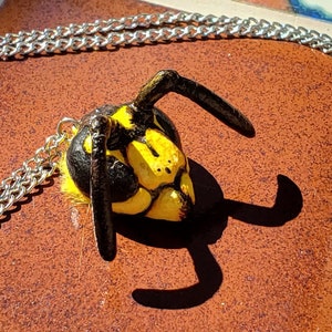 May include: A silver chain necklace with a pendant shaped like a wasp. The wasp pendant is yellow and black with detailed features. The necklace is displayed on a brown, textured surface.
