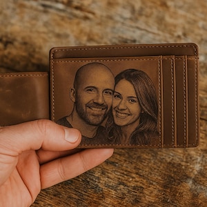 May include: A brown leather wallet with a portrait of a smiling couple engraved on the front. The wallet has card slots and a bill compartment. The leather has a natural grain and a warm, earthy tone.