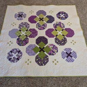 May include: A square quilt featuring a white background with a repeating pattern of octagonal shapes in shades of purple, lavender, and lime green. The quilt has a light green border and intricate white stitching throughout the fabric surface.