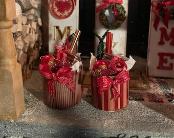 May include: Two small red and gold striped containers filled with red and gold Christmas ornaments, red ribbon, and greenery. The containers are sitting on a white surface.