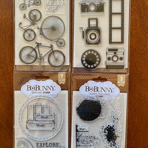 May include: Four clear plastic sheets with black and white images of bicycles, cameras, suitcases, and script textures. The sheets are labeled "Bobunny" and have the number of pieces per pack listed. The text "Life is a Beautiful Ride", "Say Cheese Stamp", "Explore Stamp", and "Script Textures Stamp" is also included.