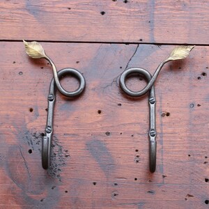 May include: Two handcrafted metal wall hooks. Each hook features a curled design with a leaf accent at the top and a hook at the bottom. The hooks are dark gray with brass-colored leaf accents, mounted on a wooden surface.