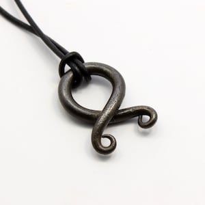 May include: A handcrafted, dark metal pendant necklace with a unique, abstract design. The pendant features a looped shape with curled ends, suspended from a black cord. The metal has a textured, aged appearance, suggesting a rustic or vintage style.