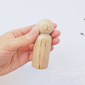 Puede incluir: Una muñeca de madera simple con una cabeza redonda y un cuerpo simple. La muñeca está sostenida en una mano.