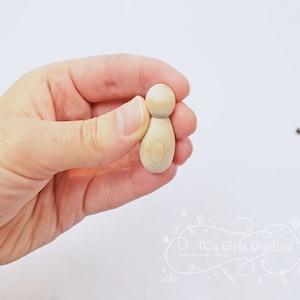 May include: A plain, unfinished wooden doll with a rounded head and body. The doll is held in a hand.