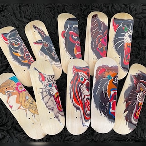 May include: A selection of miniature skateboards featuring tattoo-style artwork. The designs include a panther, tiger, bear, and a woman's portrait, all in vivid colours. The boards are made of light-coloured wood.