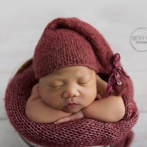 Puede incluir: Un bebé recién nacido durmiendo en una envoltura y un gorro de punto rosa. El bebé lleva un gorro de punto rosa con un lazo.