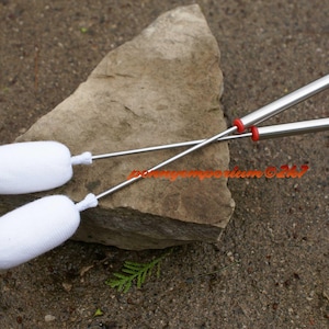 May include: Two white, soft, round-tipped drumsticks with red-tipped metal handles. The drumsticks are crossed over each other and resting on a gray rock.