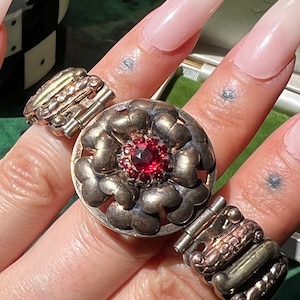 May include: A hand displaying several rings, including a gold ring with a red stone, a silver ring with a central red stone, and a silver and gold ring. A black and white checkered cup is in the background.