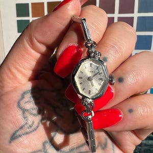 May include: Vintage Timex watch with a silver octagonal face and a silver band. The watch face has the word "TIMEX" printed on it. The watch is held up against a colorful background.
