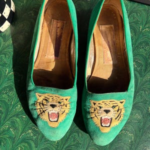 May include: A pair of bright green suede loafers featuring an embroidered tiger head on the toe of each shoe. The tiger design includes yellow, black, and red thread details. The interior of the shoes is lined with tan leather and shows signs of wear.
