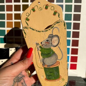 May include: A light tan wooden wall pocket featuring a hand-painted gray mouse wearing a green sweater, sitting on a green spool of thread while holding a needle. The top section is decorated with a painted garland of small green thread spools.