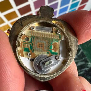 以下が含まれることがあります： 裏蓋を取り外した金色の円形時計ケース。内部の電子回路基板、中央のマイクロチップ、および小さな金属製ブラケットとネジで固定された銀色のボタン電池が見える状態。