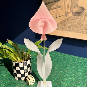 May include: A decorative, abstract flower sculpture with a pink petal and white leaves. The sculpture is on a white base. A black and white checkered pot with a plant and a die are also in the image.