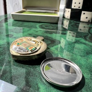 以下が含まれることがあります： 内部の電子回路基板が露出したゴールドトーンのハート型ペンダントと、取り外された円形のシルバートーンの金属製裏蓋。背景には、内側にKrementzというブランド名が印刷されたクリーム色のジュエリーボックスが置かれています。