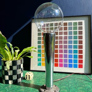 May include: A chrome-finished table lamp with a large, clear globe bulb. The lamp has a cylindrical stem and a circular base. A framed color chart and a potted plant in a black and white checkered pot are in the background. A die is also visible.