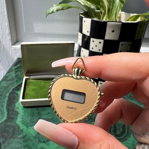 以下が含まれることがあります： デジタル表示とロープ状の縁取りが特徴の、ハート型のゴールドトーンペンダントウォッチ。文字盤の画面下部には「QUARTZ」の文字が黒でプリントされています。上部にはチェーンを通すための小さな三角形のバチカンが付いています。