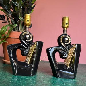 May include: A pair of black ceramic table lamps with gold accents. The lamps are in the shape of a stylized human figure with a gold circle around the neck. The lamps are sitting on a green surface.