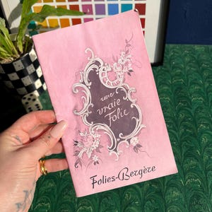May include: A pink booklet with the text "une vraie Folie" in an ornate frame, and "Folies-Bergère" below. The cover features floral and scroll designs. The booklet is held in a hand with long nails, and is set against a green patterned background.