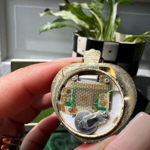 以下が含まれることがあります： リンゴの形をしたゴールドトーンのペンダント。裏面が開いており、内部の電子回路基板、小さな銀色の電池、白いプラスチック製のハウジング内に小さなネジで固定された金属部品が見える。
