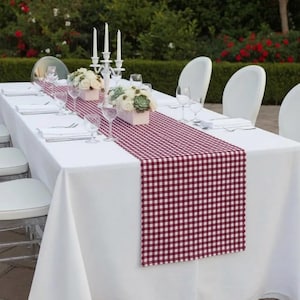 Puede incluir: Una mesa de comedor formal preparada para una reunión, con un mantel blanco y un camino de mesa a cuadros burdeos y blanco. La mesa está puesta con sillas blancas, cristalería y centros de mesa florales.