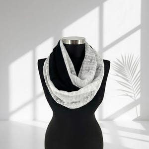 May include: A black and white infinity scarf. The scarf is draped on a black mannequin. The top layer is white lace, and the bottom layer is black fabric. The scarf is a fashionable accessory.