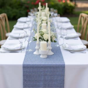 Op de afbeelding: Een prachtig gedekte eettafel in de buitenlucht met een wit tafelkleed en een blauw-wit geruite tafelloper. Witte borden, wijnglazen en kaarsen in witte houders zijn voor elke plaatsing gerangschikt. Bloemschikkingen voegen een vleugje elegantie toe.