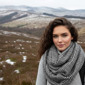 May include: A woman wearing a gray sweater and a black and white patterned scarf, posing outdoors. The background features a mountainous landscape with patches of snow. The scarf is wrapped around the neck, providing warmth.