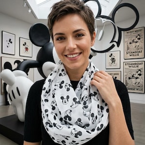 May include: A white infinity scarf featuring a repeating black and white graphic print of Mickey Mouse faces. The lightweight fabric is draped in a circular loop around the neck, showcasing a consistent pattern of the character's head in various expressions.